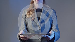 An adult woman in a gray business suit stands and holds a graphics tablet. No face