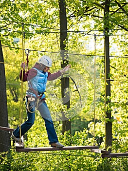 Adult woman in a helmet and with a safety system cautiously walking on suspension bridge on the background of trees