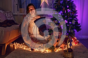 Adult woman with her son knit clothes by the Christmas tree in the eve