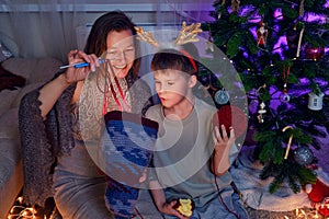 Adult woman with her son knit clothes by the Christmas tree in the eve