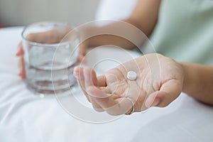 Adult woman holding pill and glass of water, taking medicine on bed in morning at home. Migraine, painkiller, headache, influenza