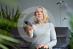 Adult woman at home sitting on the couch and watching tv, she is holding a remote control.