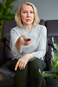 Adult woman at home sitting on the couch and watching tv, she is holding a remote control.