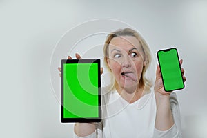 adult woman looks sideways at one of tablets in hands she holds phone and tablet with green chromakey screen cheerfully