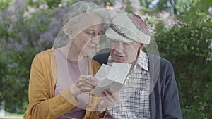 Adult woman and man looking old photos remembering happy moments sitting on a bench in the park. Mature couple in love