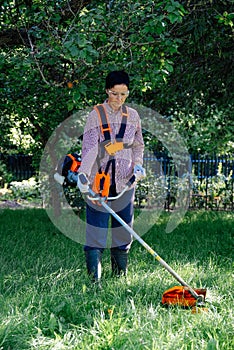 Adult woman mows the grass in the backyard using string trimmer. Garden work concept