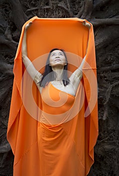 Adult woman in orange dress, dancing under tree roots