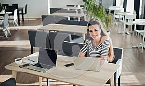 Adult woman with a perfect smile sitting in a cafe with a laptop, holding a cup of coffee and looking at the camera.