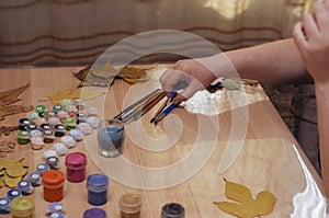 An adult woman sits at a table indoors. On the table are various paints, paint brushes, and dry tree leaves. Drawing on the fallen