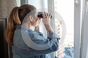 Adult woman spying out the window looking through binoculars, a domestic room