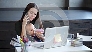Adult woman is talking by mobile phone, holding baby on a knees in a home.