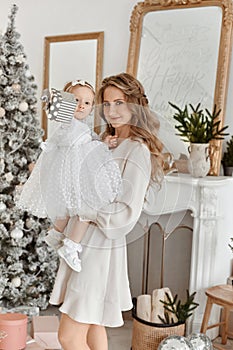 Adult woman with tender makeup and long hair posing with her cute toddler daughter in decorated Christmas interior