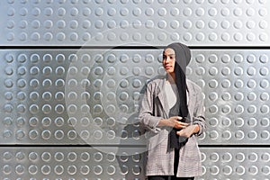 Adult woman wearing a headscarf and holding her purse standing using her phone looking away