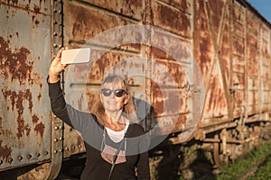 Adult woman wearing sunglasses taking a selfie with her smartphone of abandoned train cars