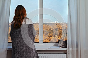 Adult woman at the window wrapped in a grey blanket next to a cat. Autumn nature outside the window and home comfort