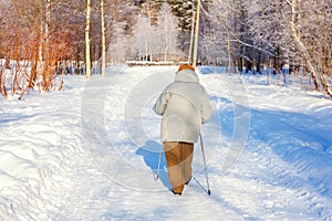 Adult woman in winter sportswear with sticks for Nordic walking on winter landscape city park background, back view