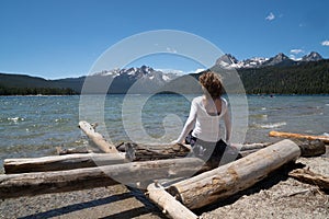 Adult woman 30-35 years sits on a pile of logs on the shoreline of Redfish Lake in Stanley Idaho in the Sawtooth Mountains in
