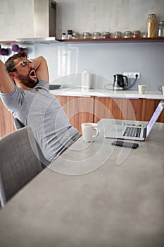 Adult young man with beard and tattoo, wearing glasses, yawns, stretching in front of laptop on table. modern lifestyle, freelance