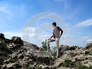 The adult young man climbed a rock and looks into the distance in a winner pose. Dark-haired slim guy in a T-shirt, white pants