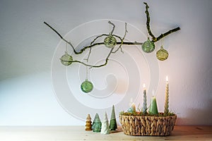 Advent and Christmas decoration with four different candles in a basket and small artificial trees under a hanging bare branch