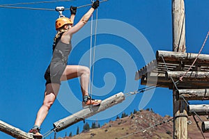 Adventure climbing rope park - a young woman walks along logs and ropes at a height against the background of mountains and blue s