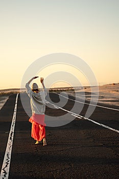 Adventure concept. Back view of a hippie woman walking alone and barefoot on the highway, dancing