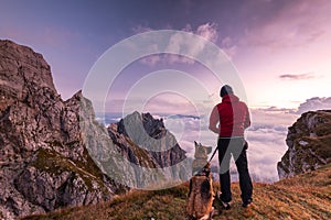 Adventure Man with Dog at High Mountains Peak at Sunrise. Togetherness and Friendship Concept