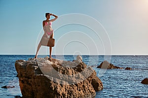 Adventure trip idea. Lady in pink bikini with suitcase standing on rock. Summer holiday. Time for travel. Beach vacation