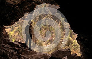 ADVENTURER WITH HAT AND BACKPACK IN A CAVE IN FRONT OF A PINE FOREST