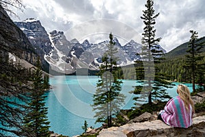 Adventurous blonde woman sits on the edge of a cliff along the rockpile trail at Moraine Lake, viewing the Canadian Rockies