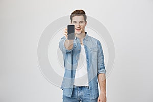 Advertisement concept. Handsome young man holding smartphone, showing gadget to camera, isolated over white background