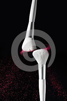 Freeze motion of two white makeup brushes with falling pink powder, black background. Advertising photo.