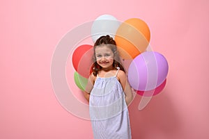 Advertising photography with cute baby girl in summer dress holding colorful balloons, posing against pink background, copy space