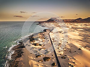 Aerial above view of yellow tropical sandy beach with black long way road and car traveling - ocean blue waves and shore - sunset