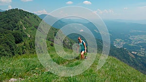 AERIAL: Active girl and her dog hike up grassy mountain overlooking green valley
