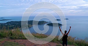 Aerial: Athletic young man stands with his hands up on the edge of the mountain.