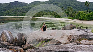 Aerial circle footage around blonde tanned girl sitting in yoga pose and meditating on granite rock against exotic