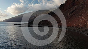 Aerial dolly close to cliff and boulders toward beach with black sand