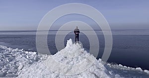 Aerial dron view of young active happy man staying on the ice glaciers near coastline of winter sea. Drone flies into