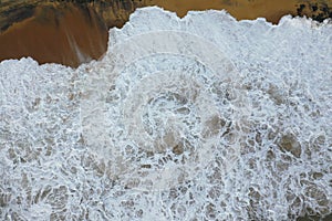 Aerial drone bird view shot of the sea shore with yellow sand, black rocks, large white waves and foam crashing on the beach