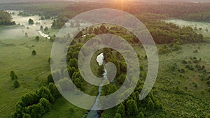 Aerial drone flies over winding river flows through green meadow and forest. Fog over water surface