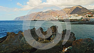 Aerial drone footage of rocky volcanic shoreline deep blue ocean water with cityscape on background. Tenerife, Atlantic