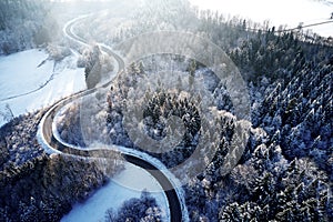 Aerial drone view of a curved winding road through the forest up in the mountains in the winter with snow covered trees