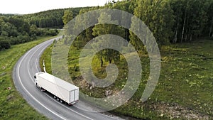 Aerial drone view of a truck car driving on a bending road among green trees. Scene. White long cargo car on the empty