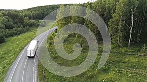 Aerial drone view of a truck car driving on a bending road among green trees. Scene. White long cargo car on the empty