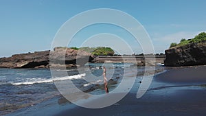 Aerial drone view of woman with sexy butt in black swimsuit go into the ocean with foam waves on black sand beach with