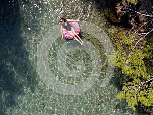 Aerial drone view of young woman with open arms in swimsuit lying and relaxing on big pink donut float swims on water