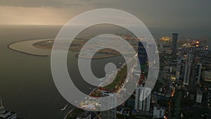 Aerial evening shot of modern urban district at sea coast. Large city after sunset. Illuminated cranes in seaport in