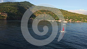 Aerial flyby around scarlet red sail boat floating on quiet ocean water near sea shore with green hills and volcano in