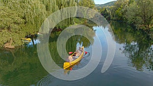 AERIAL: Flying above a group of friends paddling a large canoe along calm river.